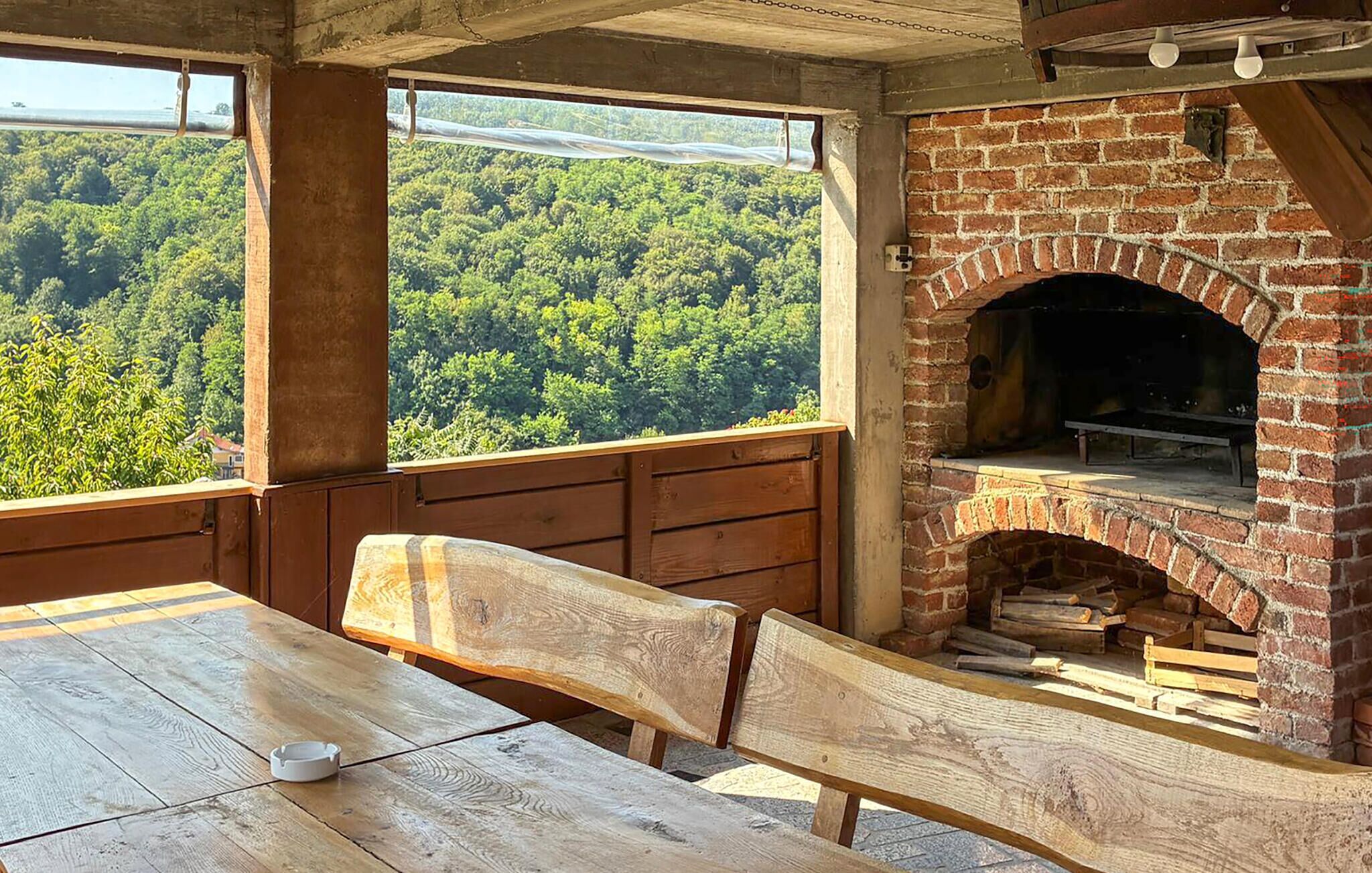 A rustic outdoor seating area with a brick fireplace, wooden benches, and a view of a forested landscape.