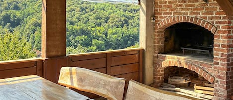 A rustic outdoor seating area with a brick fireplace, wooden benches, and a view of a forested landscape.