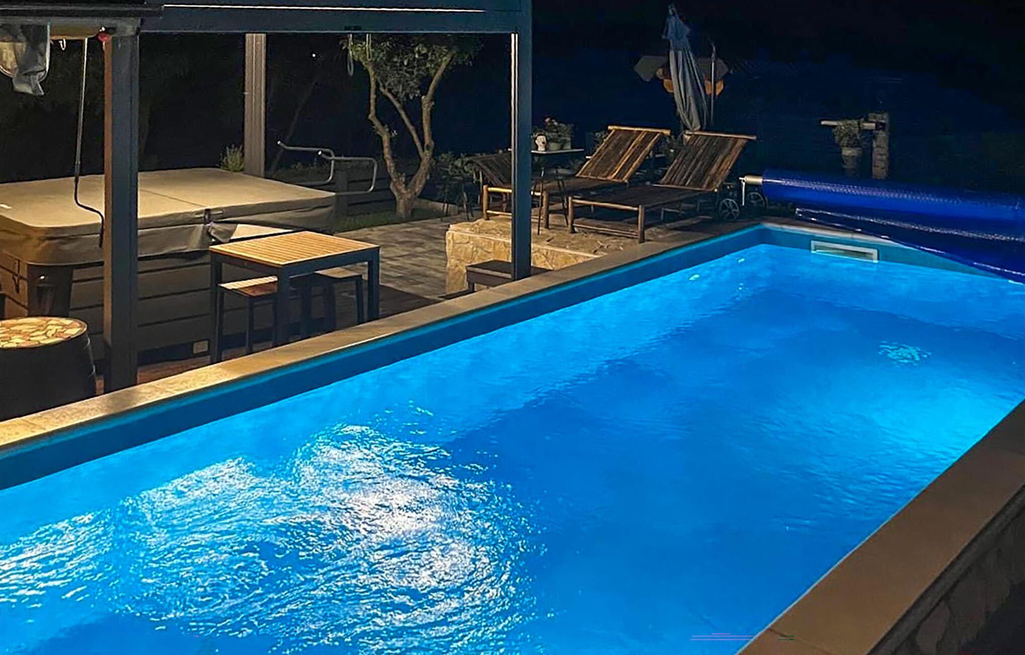 A swimming pool with lounge chairs and tables, surrounded by a covered area with a view of trees.