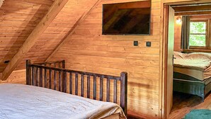 Eine Holzhütte mit geneigtem Dach, einem Bett mit Holz Kopfteil, einem an der Wand befestigten Fernseher und einem Fenster mit Blick auf Grünflächen.