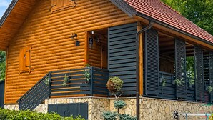 Eine Blockhütte mit Steinfundament, einem roten Ziegeldach und einer Holztreppe, die zu einer Veranda mit schwarzen Fensterläden und Topfpflanzen führt.