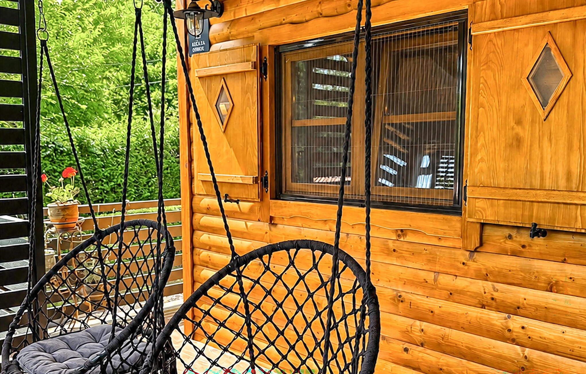 A wooden cabin with a porch, two wicker chairs, a window with a metal grill, and a door with a sign.