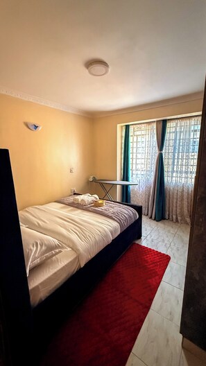 A bedroom with a bed, a red rug, a small table, and a window with curtains. - Couch Potato Pad - Tripple R Ruaka (Ruaka)