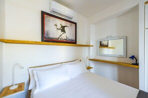 Ein Hotelzimmer mit einem Bett, einem Nachttisch mit Lampe, einer Wand-Klimaanlage, einem gerahmten Bild, einem Spiegel und einem Regal mit Lampe.