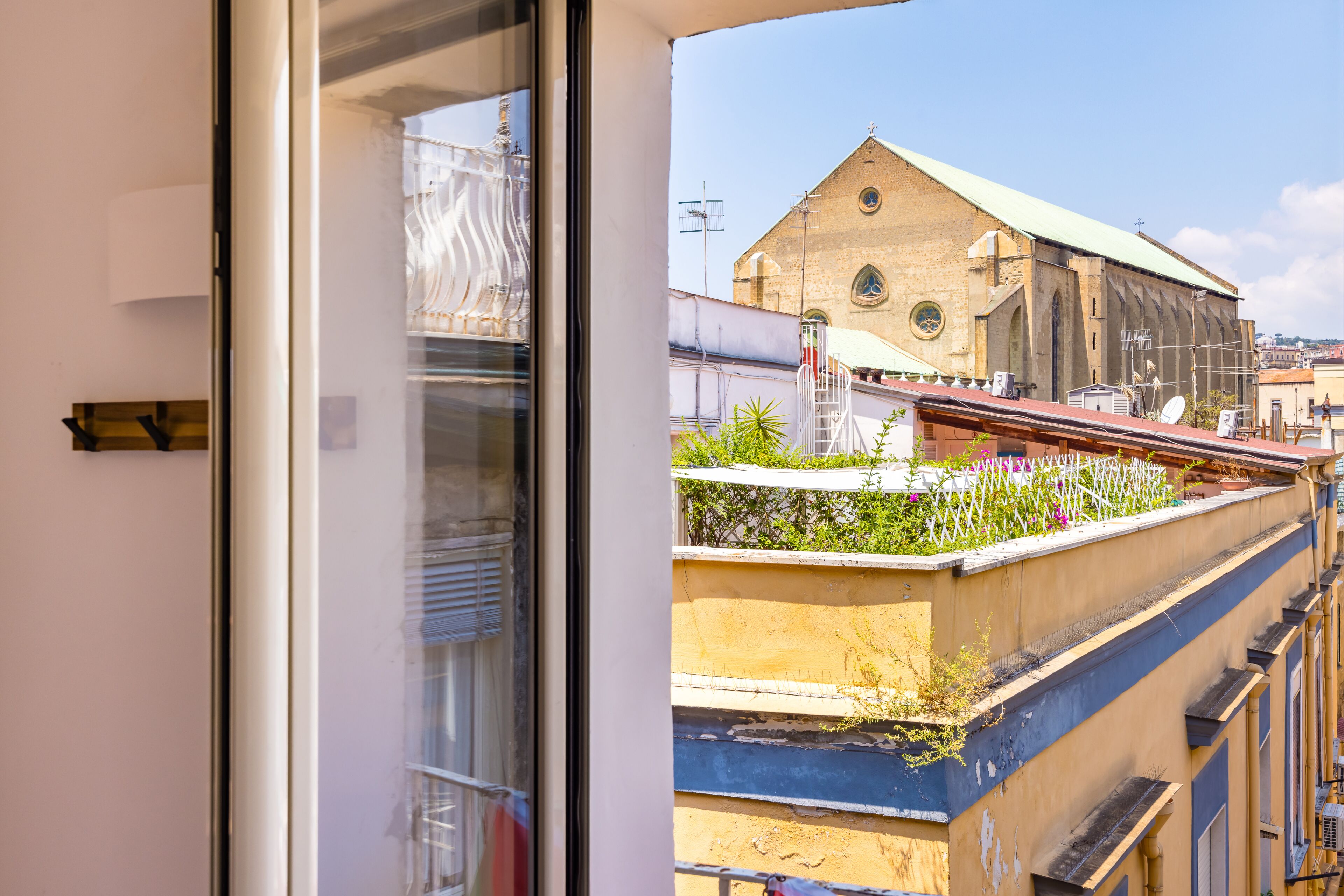 Une vue à travers la fenêtre d’un immeuble doté d’un balcon, d’une église surmontée d’un toit végétalisé et d’un ciel dégagé.