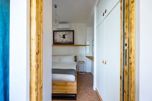 Ein schmaler Flur führt zu einem Schlafzimmer mit einem Bett, einem Nachttisch und einem gerahmten Kunstwerk an der Wand.