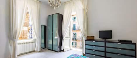 A bedroom with a bed, a dresser, a television, and a view of buildings through the window.