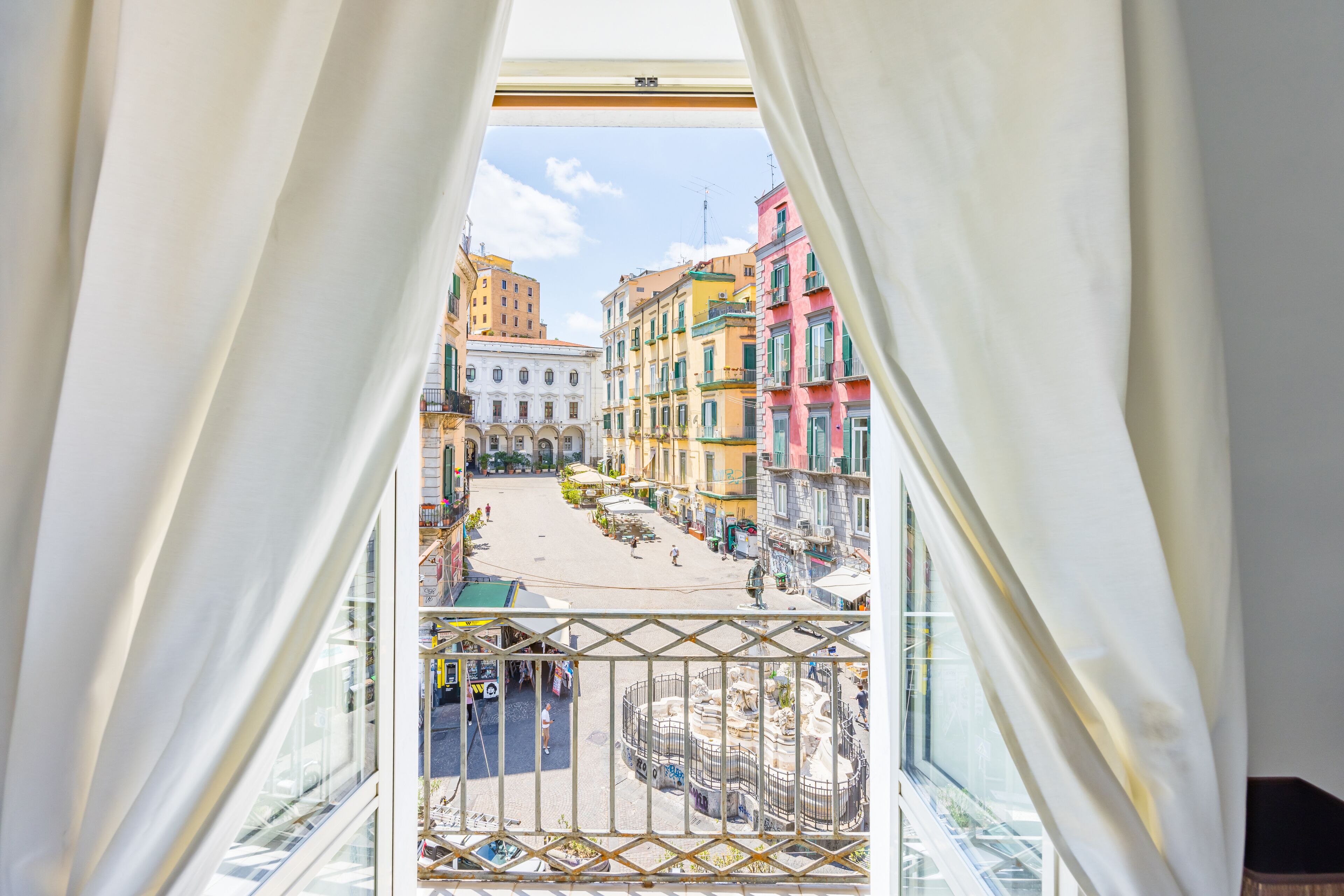 A view from a balcony with white curtains, overlooking a street with colorful buildings and a fountain.