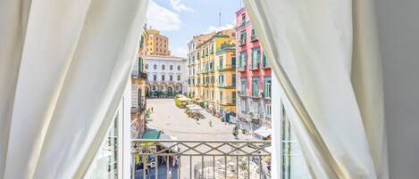 A view from a balcony with white curtains, overlooking a street with colorful buildings and a fountain.