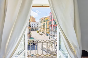 Blick von einem Balkon mit weißen Vorhängen auf eine Straße mit farbenfrohen Gebäuden und einem Brunnen.