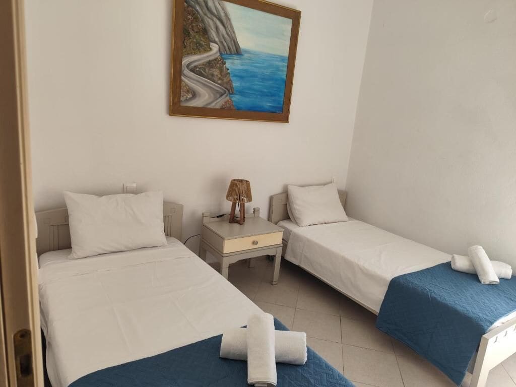 A hotel room with two single beds, a nightstand, a painting of a coastal scene, and a blue bedspread.