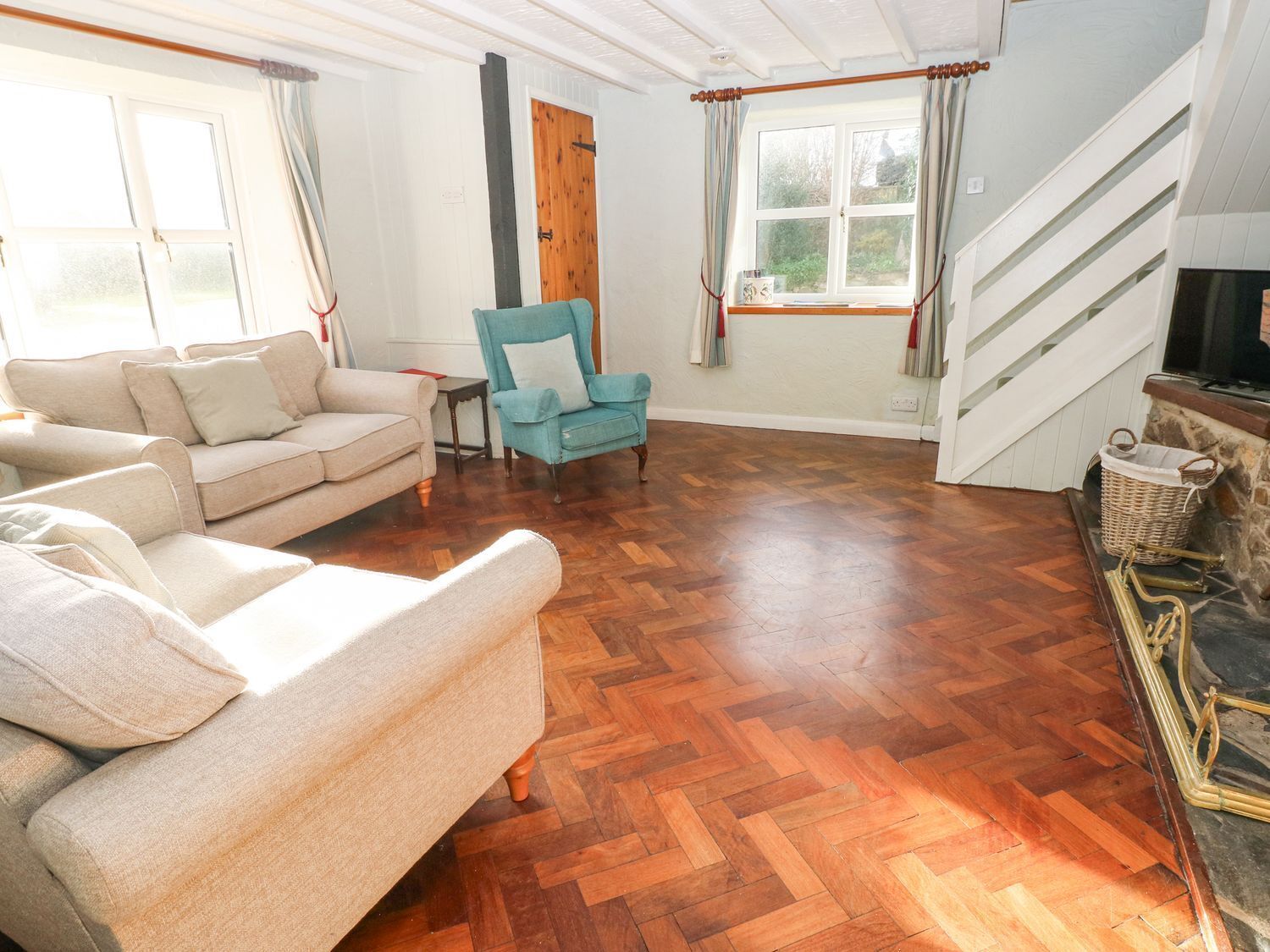 A spacious living room with a herringbone wooden floor, a beige sofa, a blue armchair, and a fireplace.