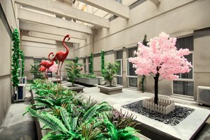 A spacious indoor atrium with a central garden area featuring a pink flowering tree, surrounded by various potted plants and a decorative flamingo sculpture.