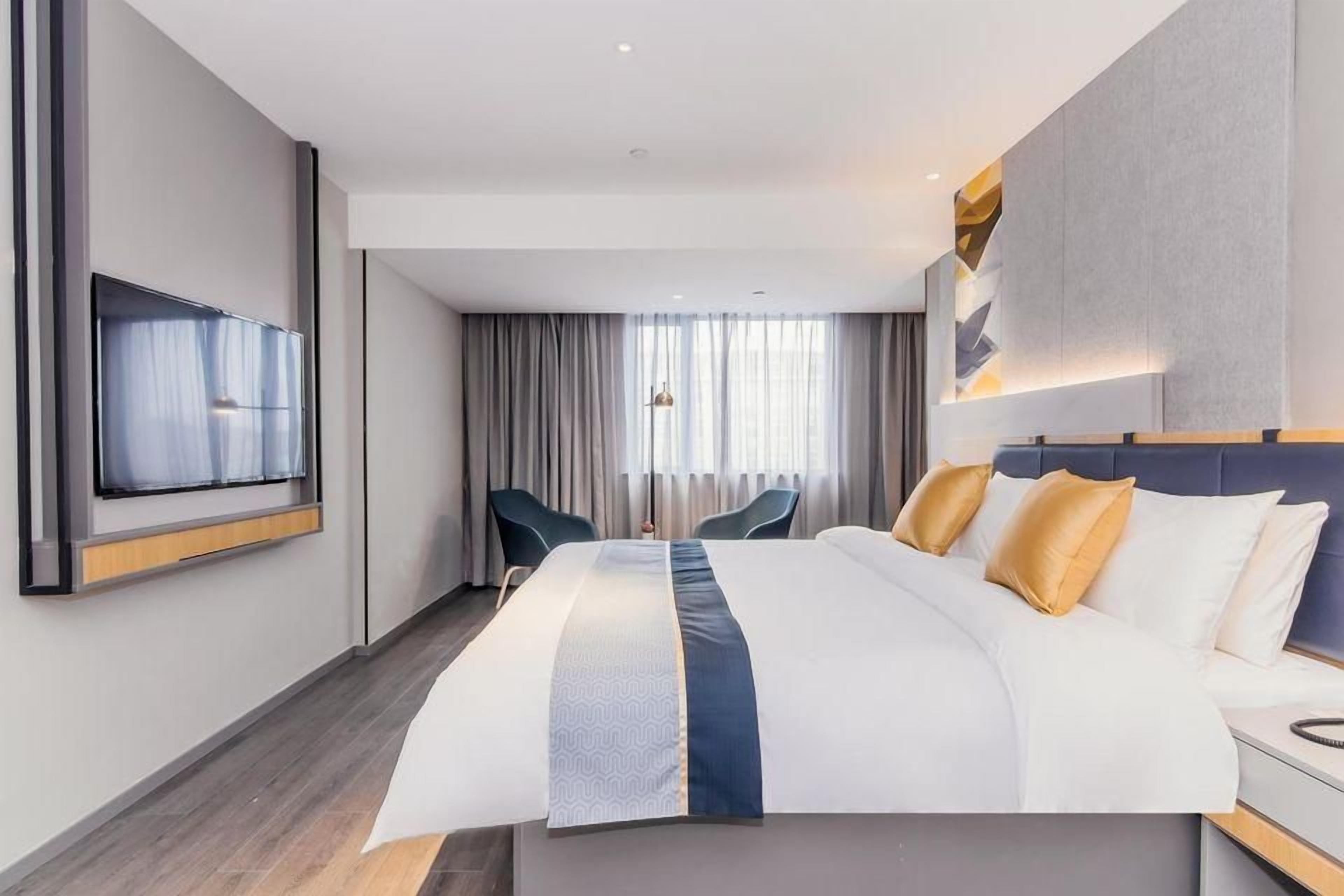 A modern hotel room with a large bed, a flat-screen TV mounted on the wall, a desk with a chair, and a view of the city through the window.