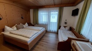 Ein Zimmer mit einem großen Bett, einem kleineren Bett und einer Bank. Vorhänge und ein an der Wand montierter Fernseher sind ebenfalls vorhanden.