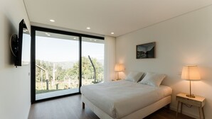 Ein Schlafzimmer mit einem großen Bett, zwei Nachttischlampen, einem Bild an der Wand und einer Glastür, die auf einen Balkon mit Blick ins Grüne führt.