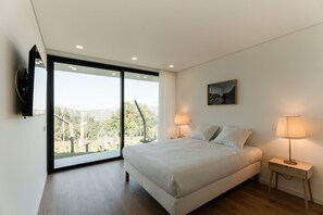 A bedroom with a large bed, two bedside lamps, a painting on the wall, and a sliding glass door leading to a balcony with a view of greenery. - Feel Discovery Douro Living Villa I (Paços de Gaiolo)