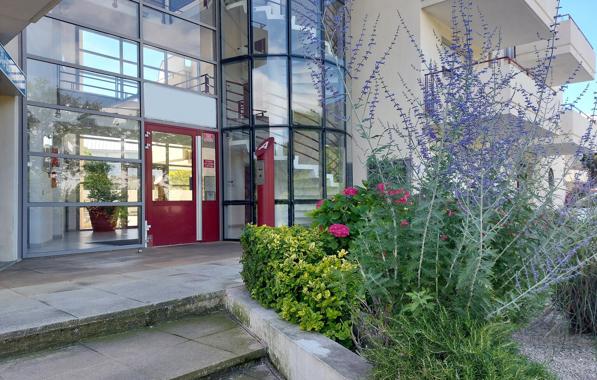 L’entrée d’un immeuble avec une porte rouge, des fenêtres en verre et un petit jardin agrémenté de diverses plantes et fleurs.