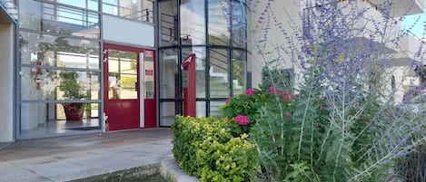 L’entrée d’un immeuble avec une porte rouge, des fenêtres en verre et un petit jardin agrémenté de diverses plantes et fleurs.