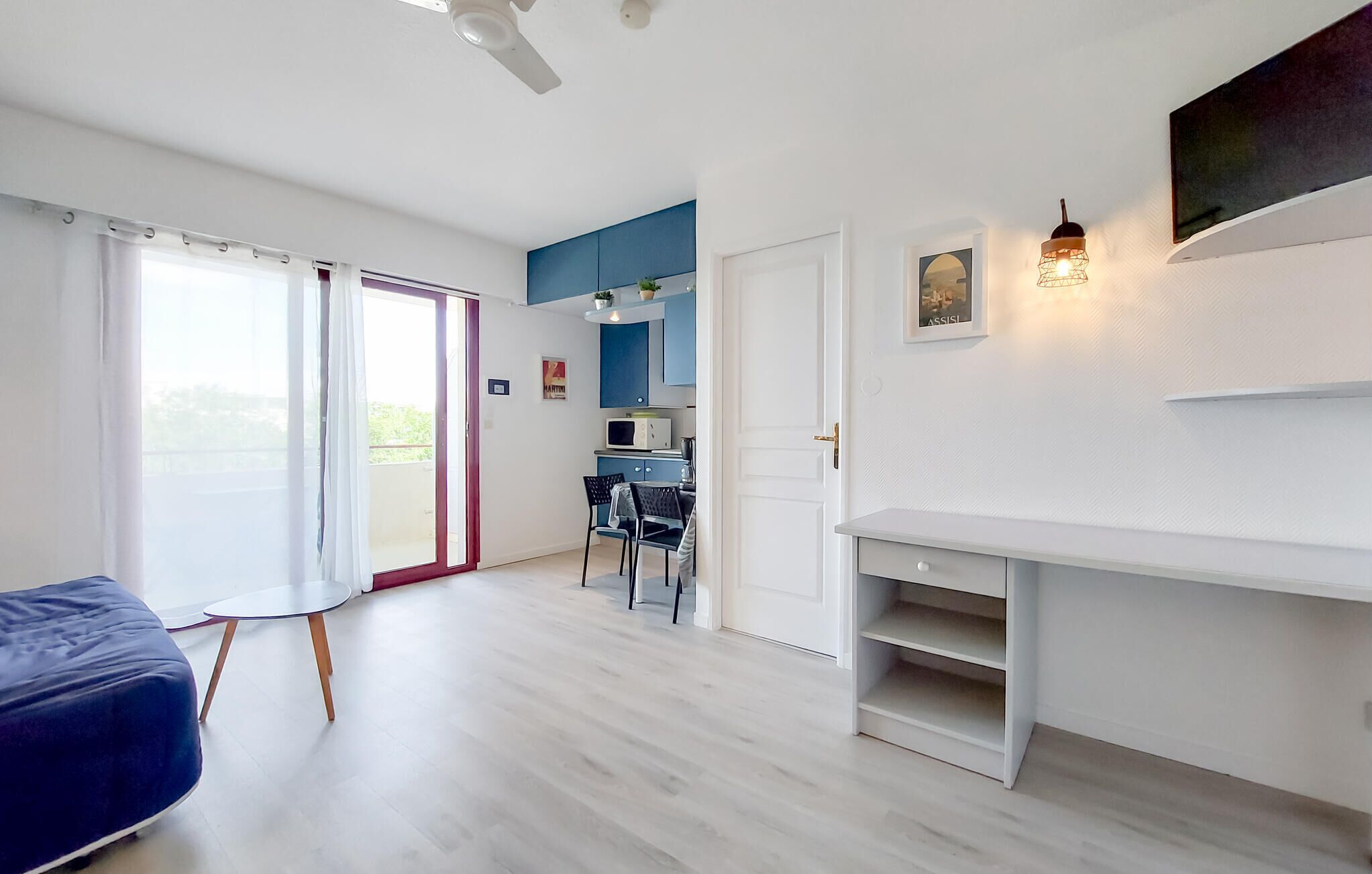 A modern living room with a sofa, a dining area with chairs, a kitchen with cabinets, and a sliding glass door leading to a balcony.