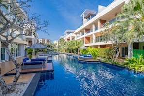 Una piscina con sillones de descanso y sombrillas, rodeada de edificios modernos y plantas tropicales.