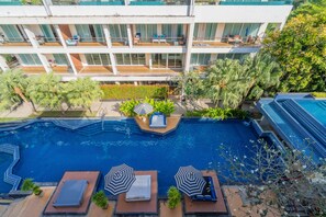 A rooftop swimming pool with lounge chairs and umbrellas, surrounded by greenery and a multi-story building.