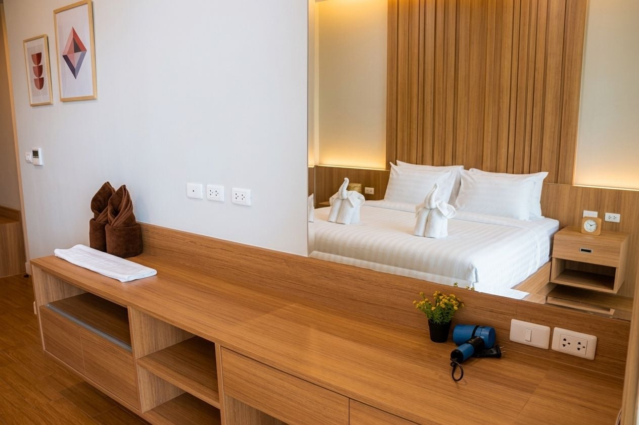 A modern hotel room with a wooden desk, a bed with white linens, a bedside table with a clock, and a wall-mounted mirror.