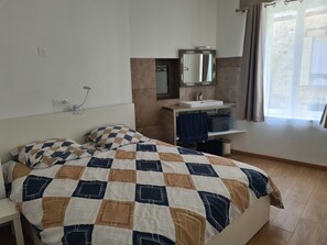 A neatly made bed with a checkered bedspread, a bedside table, a mirror, a small sink, and a window with curtains. - Vrbo Property (Thorigny-sur-Marne)