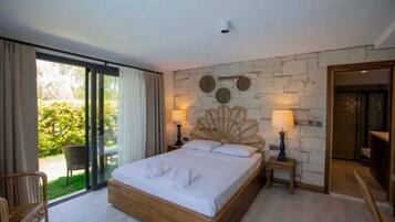 Habitación de hotel con una cama grande, puerta de cristal corredera a un jardín, pared de piedra con platos decorativos y una mesita de noche con lámpara.