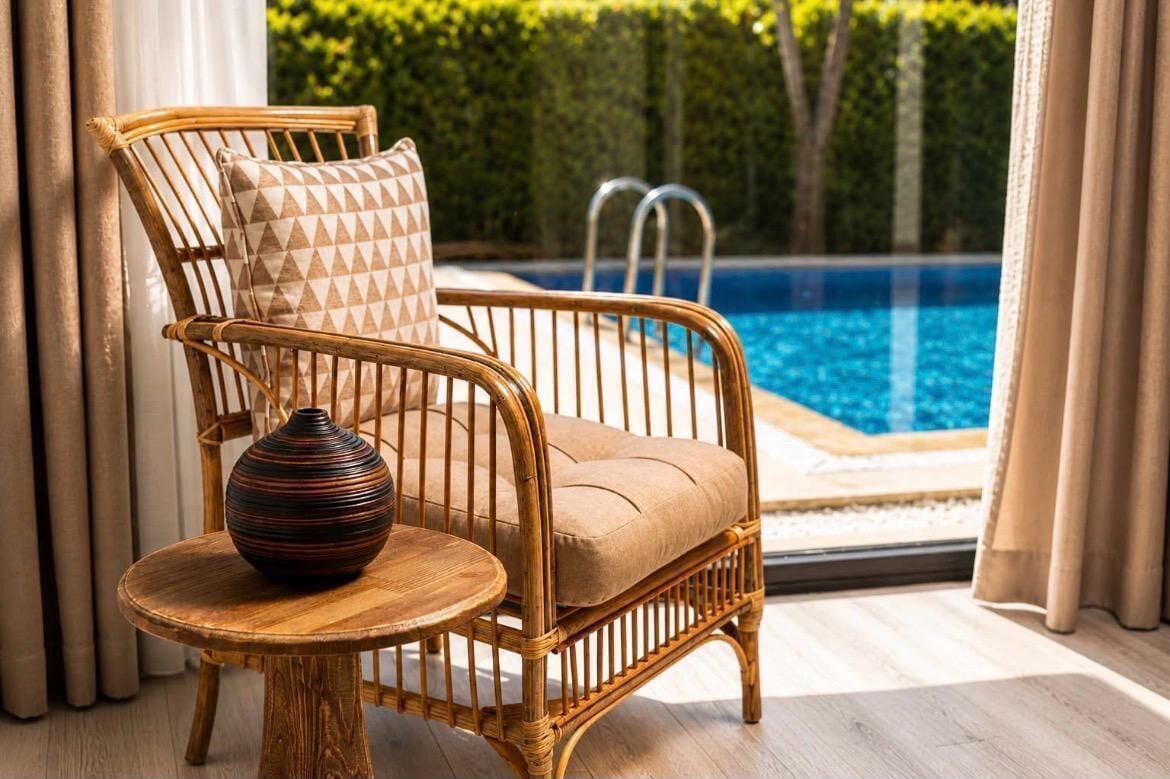 A wicker chair with a cushion and a vase on a wooden table, near a pool with a view of greenery.