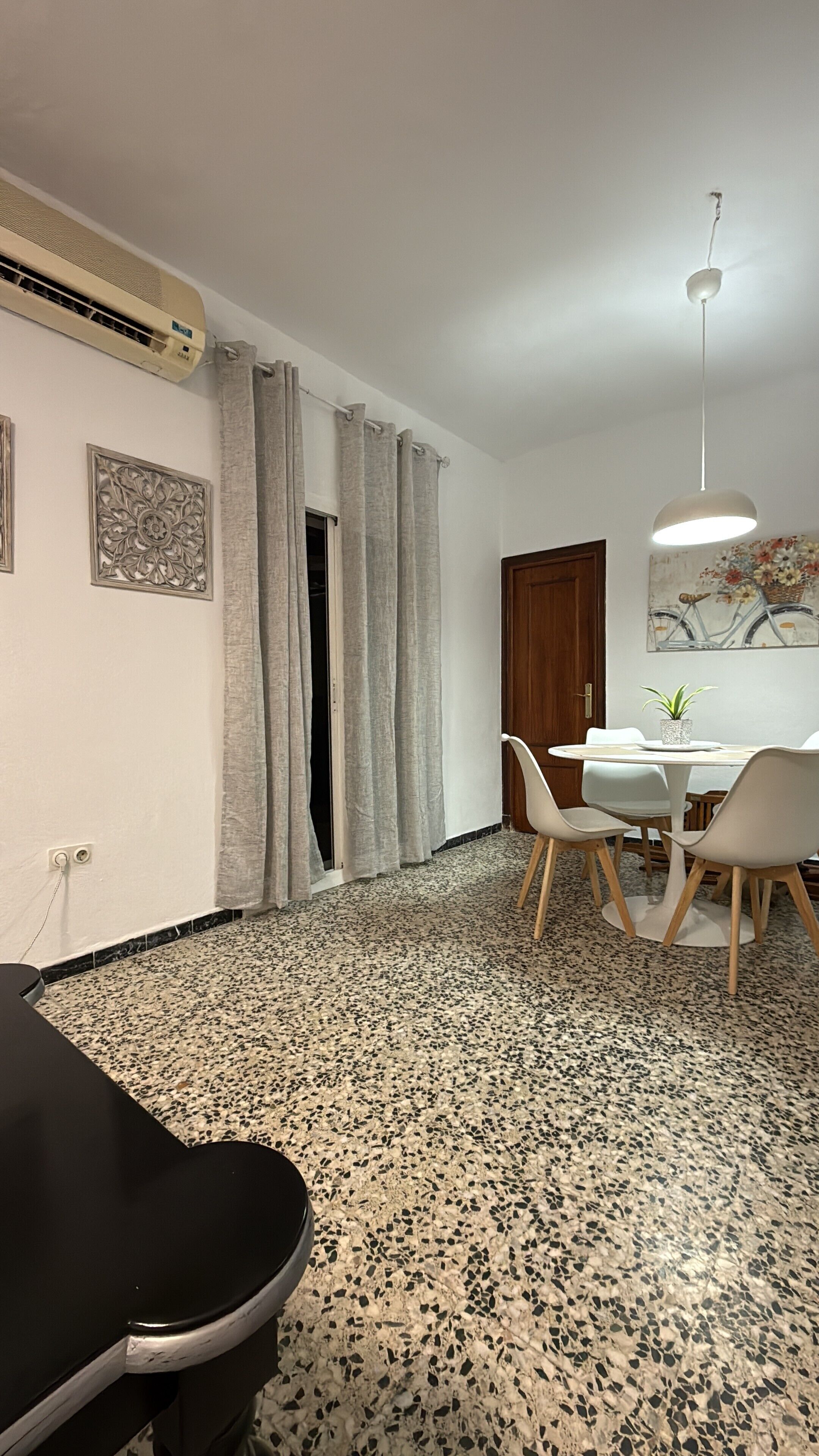 A room with a dining table, chairs, a ceiling fan, a wall-mounted air conditioner, and a door with a curtain.
