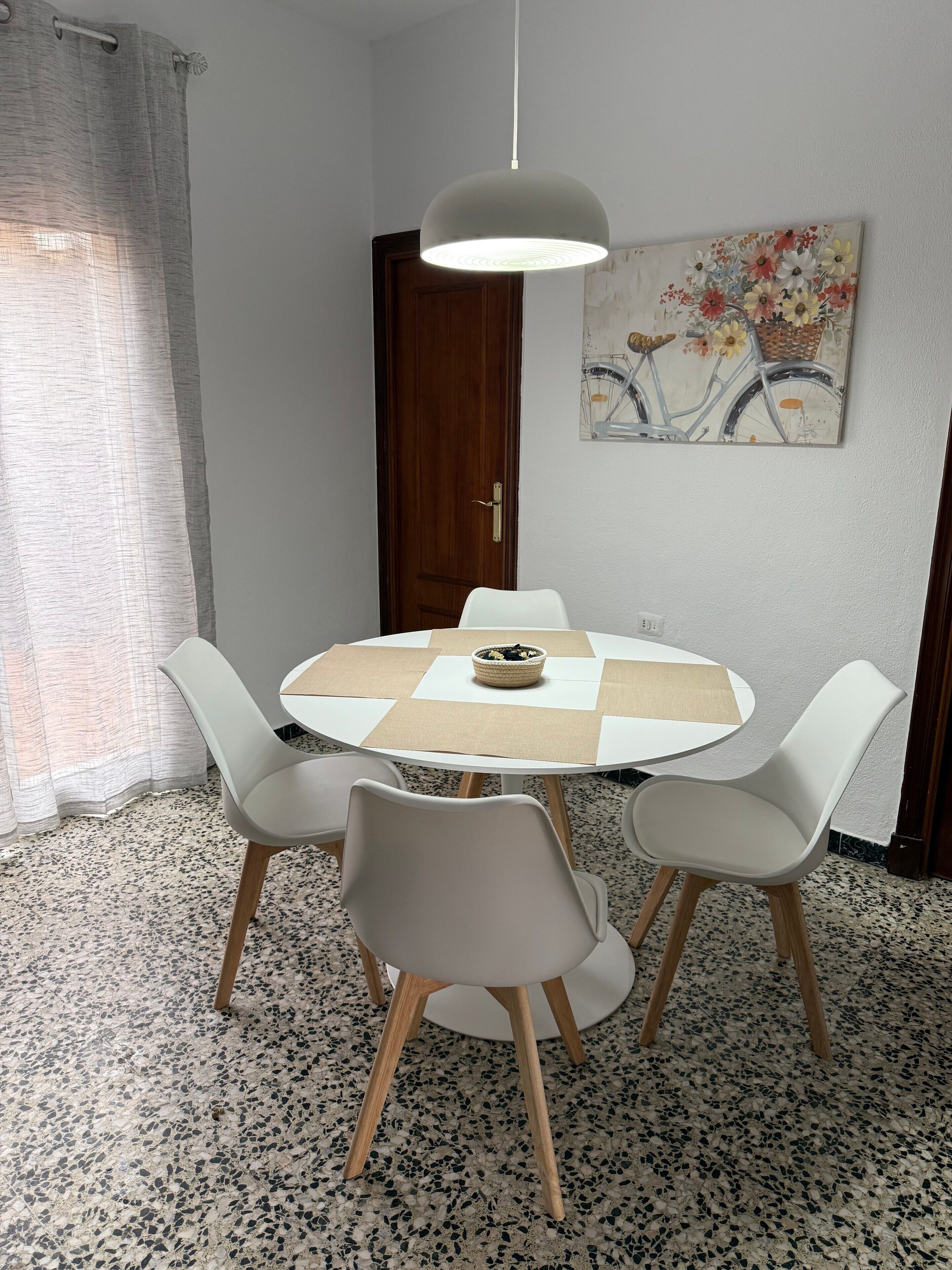 A dining area with a round table, four chairs, a hanging light fixture, a door, and a wall-mounted painting.