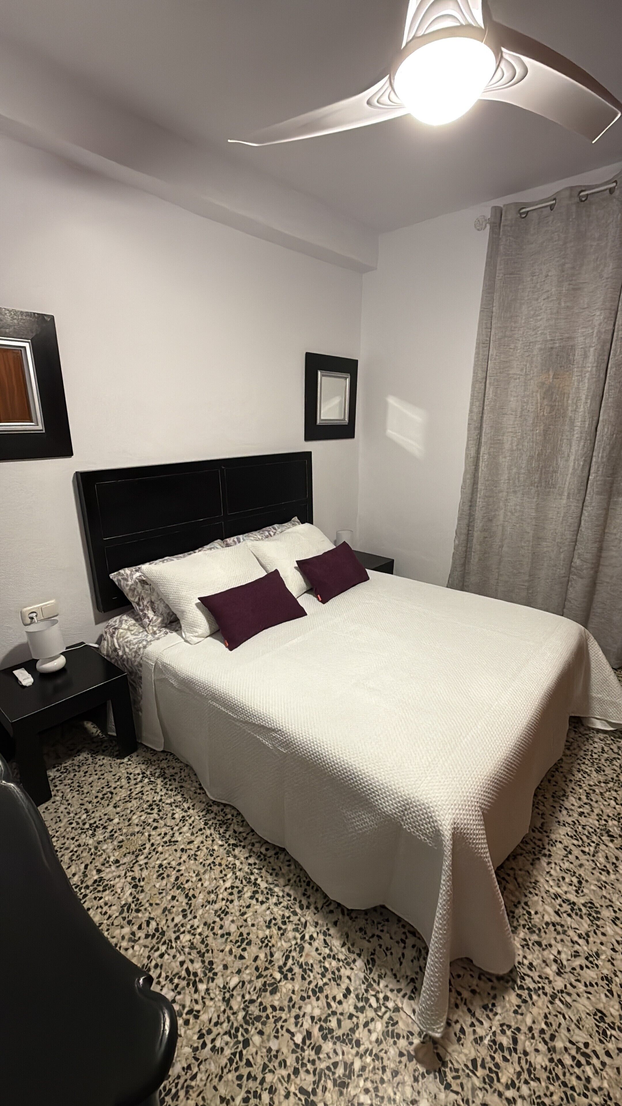 A neatly made bed with a white bedspread and two pillows, a black nightstand with a lamp, and a grey curtain.