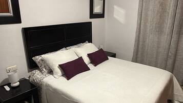A neatly made bed with a white bedspread and two pillows, a black nightstand with a lamp, and a grey curtain.