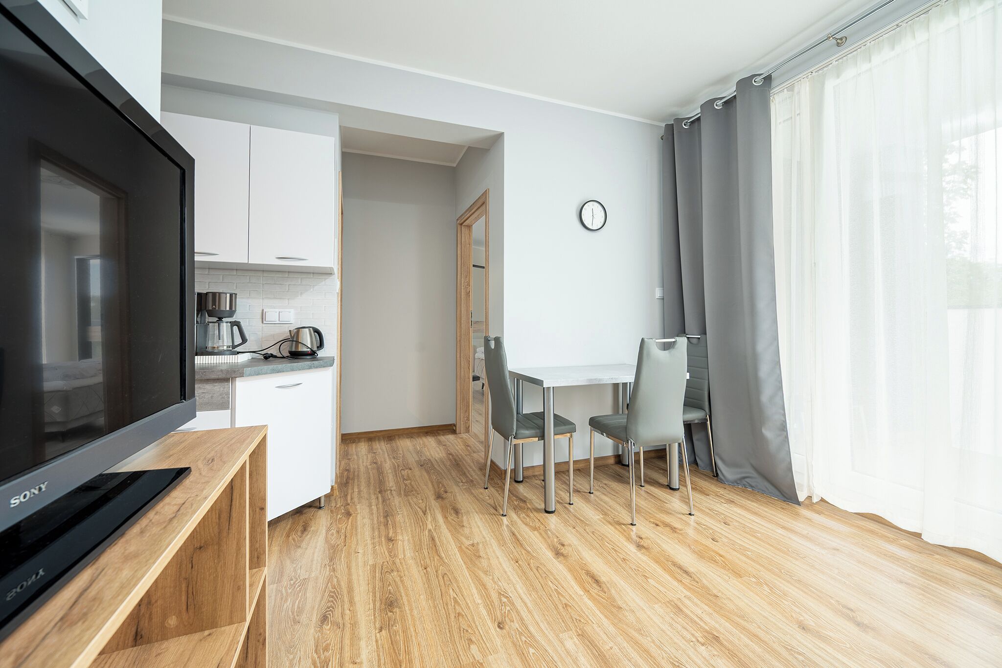 Uma sala de estar moderna com televisão de tela plana, mesa de jantar com cadeiras, cozinha com armários brancos e uma janela grande com cortinas.