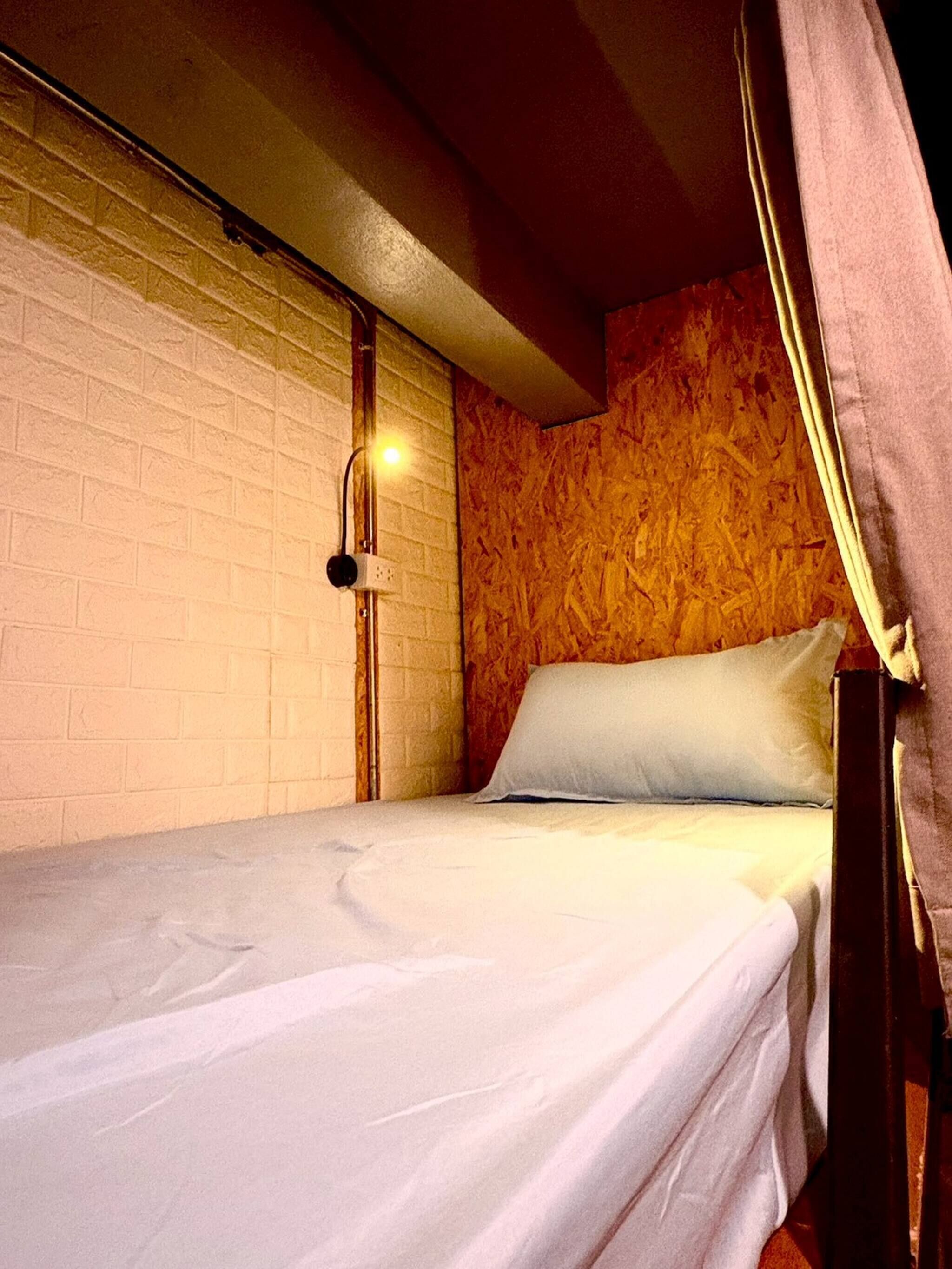 A single bed with a white mattress and a pillow, a wall-mounted light, and a wooden textured wall.
