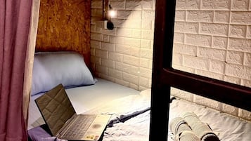 A single bed with a laptop, a pillow, and a rolled-up blanket in a room with a brick-patterned wall and a window.