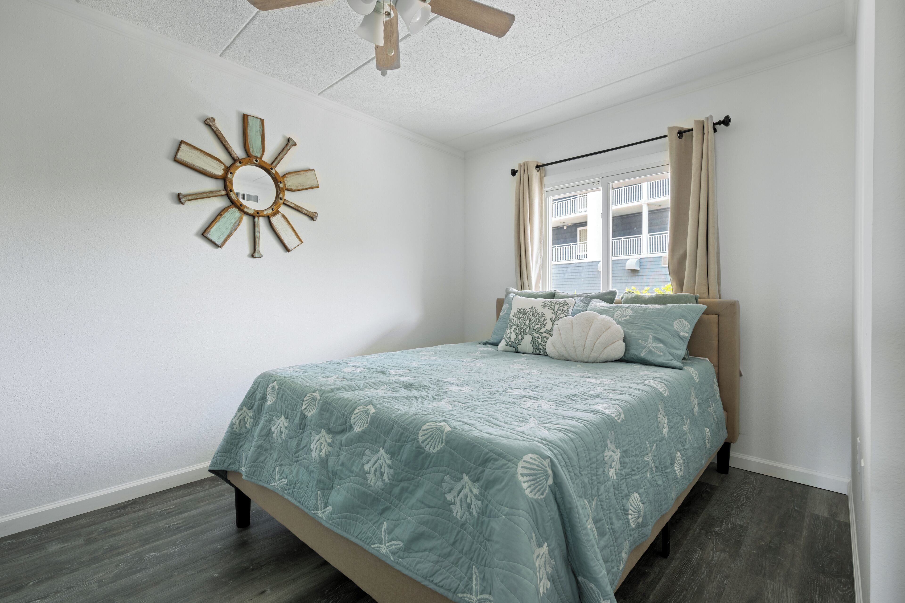 Une chambre à coucher comprenant un lit, un ventilateur de plafond, une fenêtre avec des rideaux et un miroir décoratif fixé au mur.