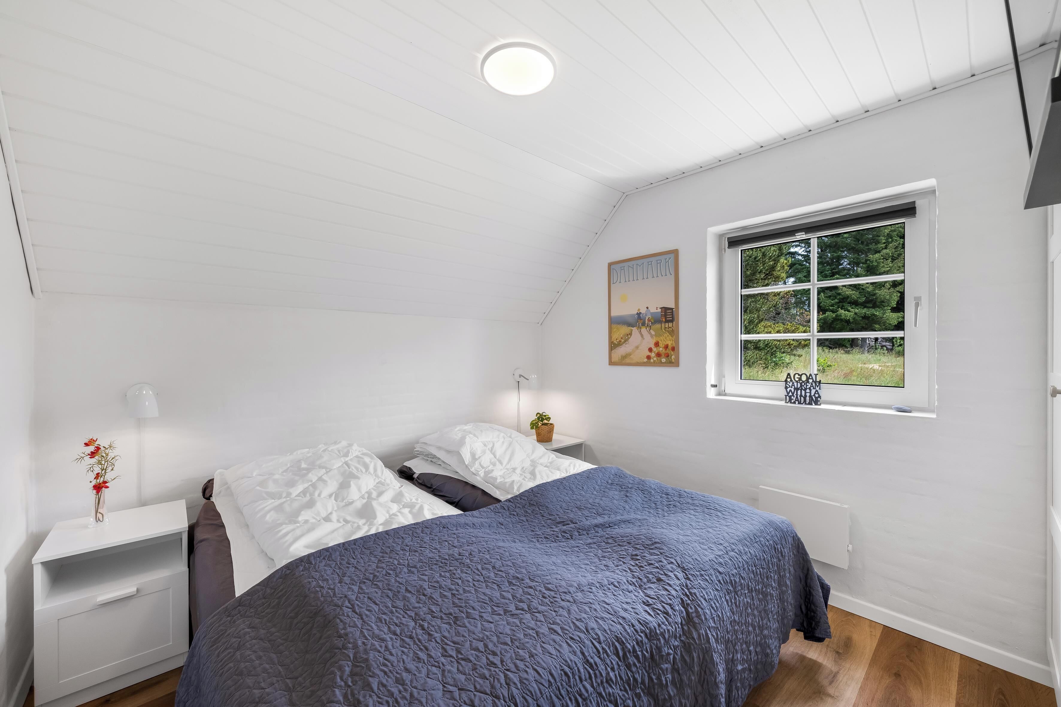 Ein Schlafzimmer mit einem Bett, einem Nachttisch, einer Lampe, einem Fenster mit Blick und einem Bild an der Wand.