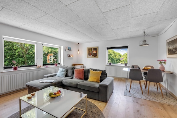 A modern living room with a sectional sofa, a dining table with chairs, and large windows with a view of greenery.