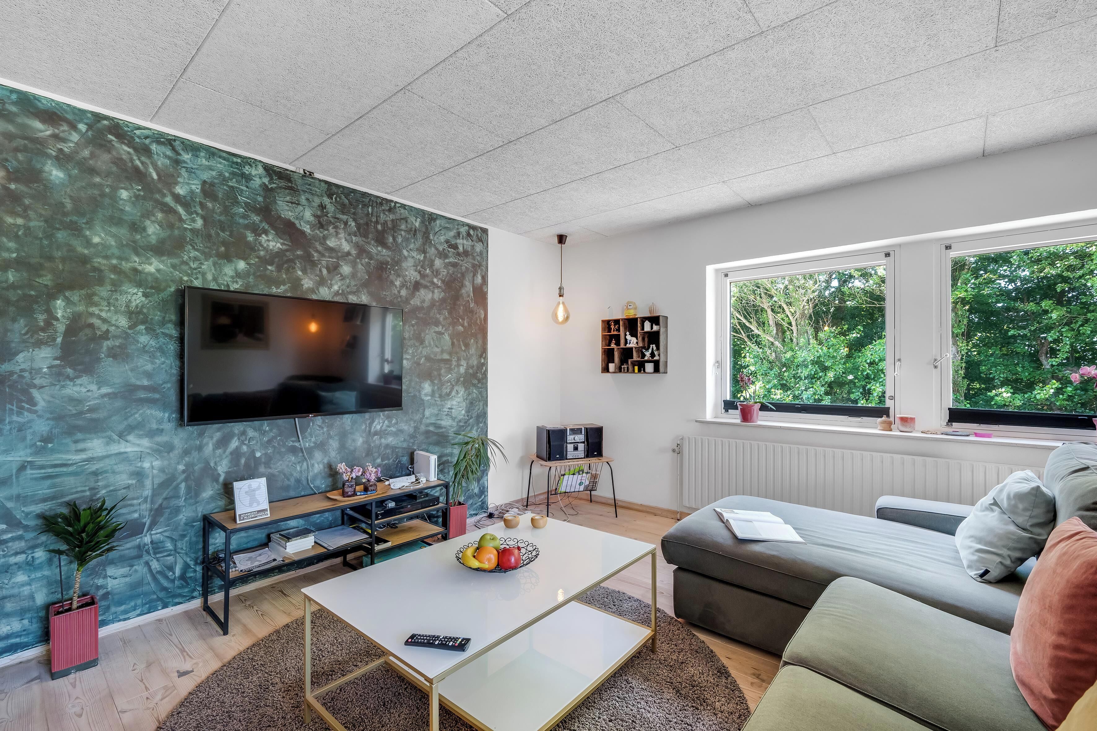 Una sala de estar moderna con una televisión de pantalla plana, un sofá, una mesa de centro y un ventanal con vista a zonas verdes.