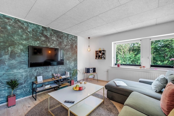 Una sala de estar moderna con una televisión de pantalla plana, un sofá, una mesa de centro y un ventanal con vista a zonas verdes.