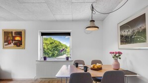 Ein Essbereich mit Tisch, Stühlen, einem Fenster mit Blick auf Grünflächen, einer Pendelleuchte und einem gerahmten Gemälde.