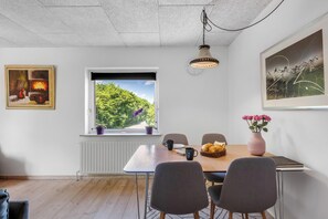Ein Essbereich mit Tisch, Stühlen, einem Fenster mit Blick auf Grünflächen, einer Pendelleuchte und einem gerahmten Gemälde.
