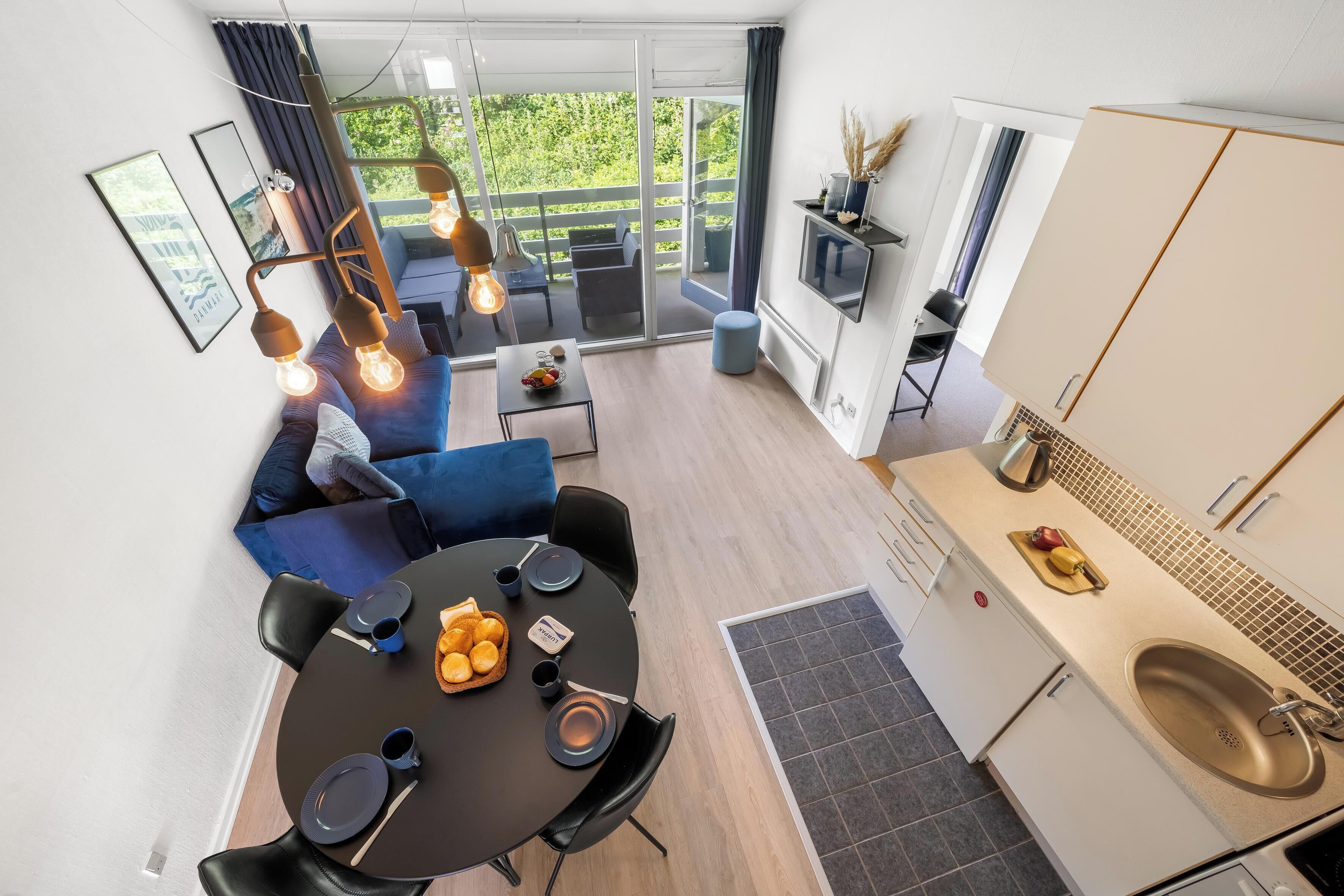 Une cuisine moderne équipée d’un évier, un coin repas avec une table dressée pour deux personnes, et un balcon agrémenté d’un canapé et de fauteuils.