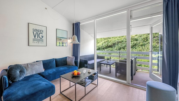 A modern living room with a blue sofa, a glass coffee table, and a view of a balcony with outdoor furniture. - Lovely holiday apartment in the heart of Søndervig close to the North Sea (Ringkøbing)