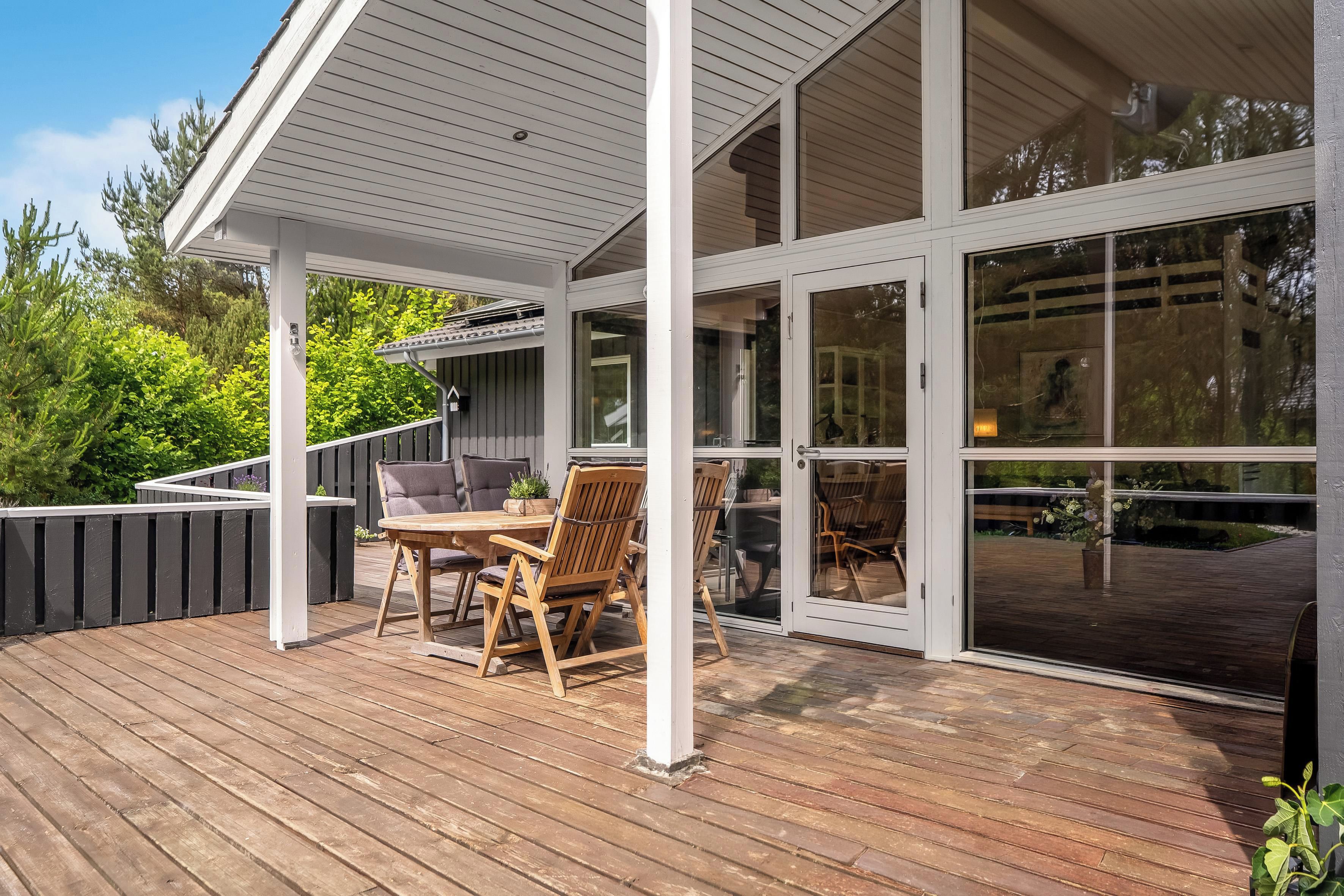 A covered outdoor deck with a table and chairs, large sliding glass doors, and a view of the surrounding greenery.