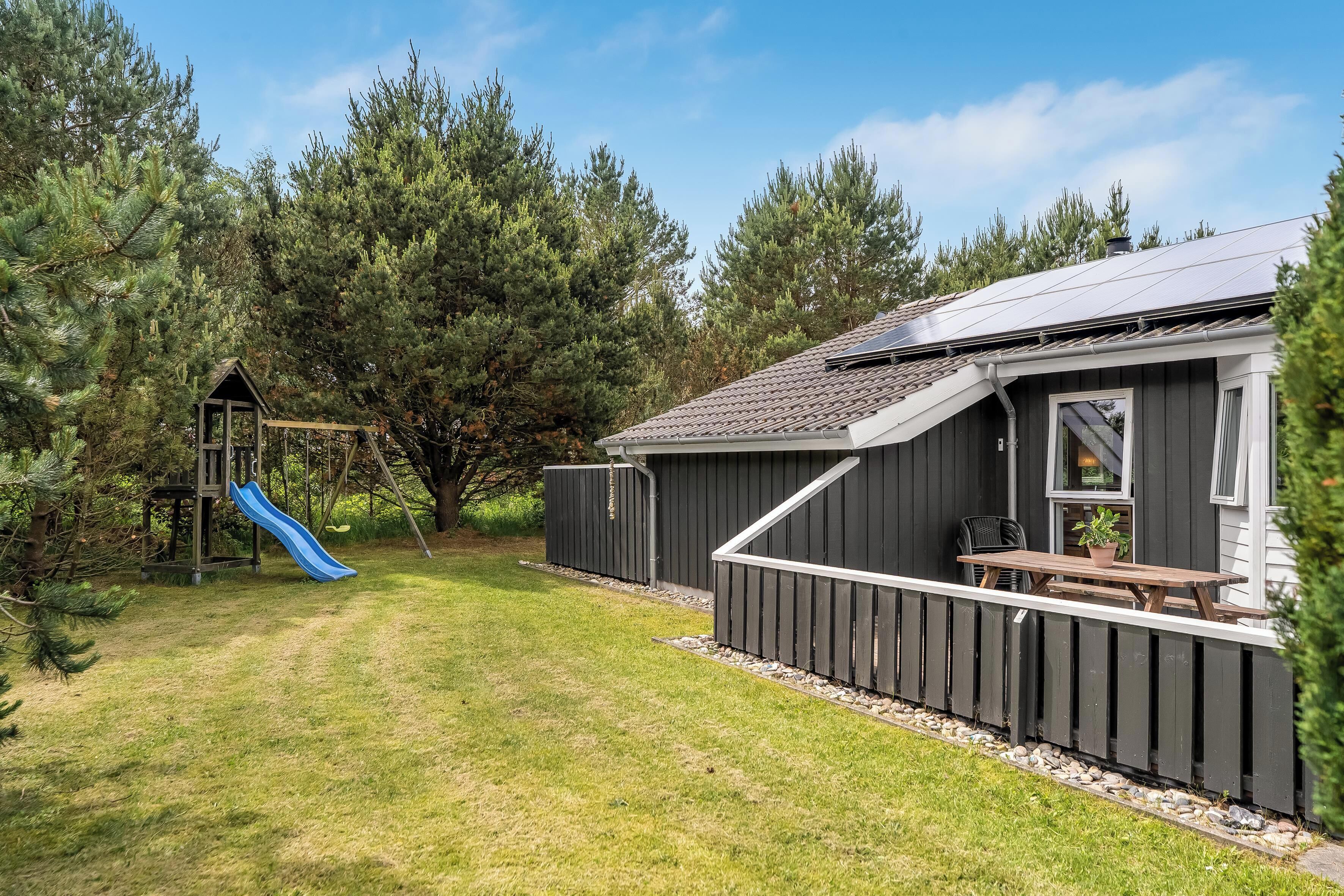 A wooden deck with a picnic table, a play structure with a slide, and a building with a sloped roof.