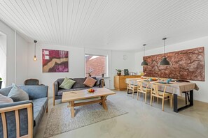 A modern living room with a sofa, coffee table, dining table, and chairs. There is a window with a view of a brick building. - Modernized ground floor holiday apartment in Stadil (Tim)