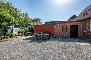 A gravel patio with a table and chairs, a brick building with a door, and a tree in the background. - Modernized ground floor holiday apartment in Stadil (Tim)
